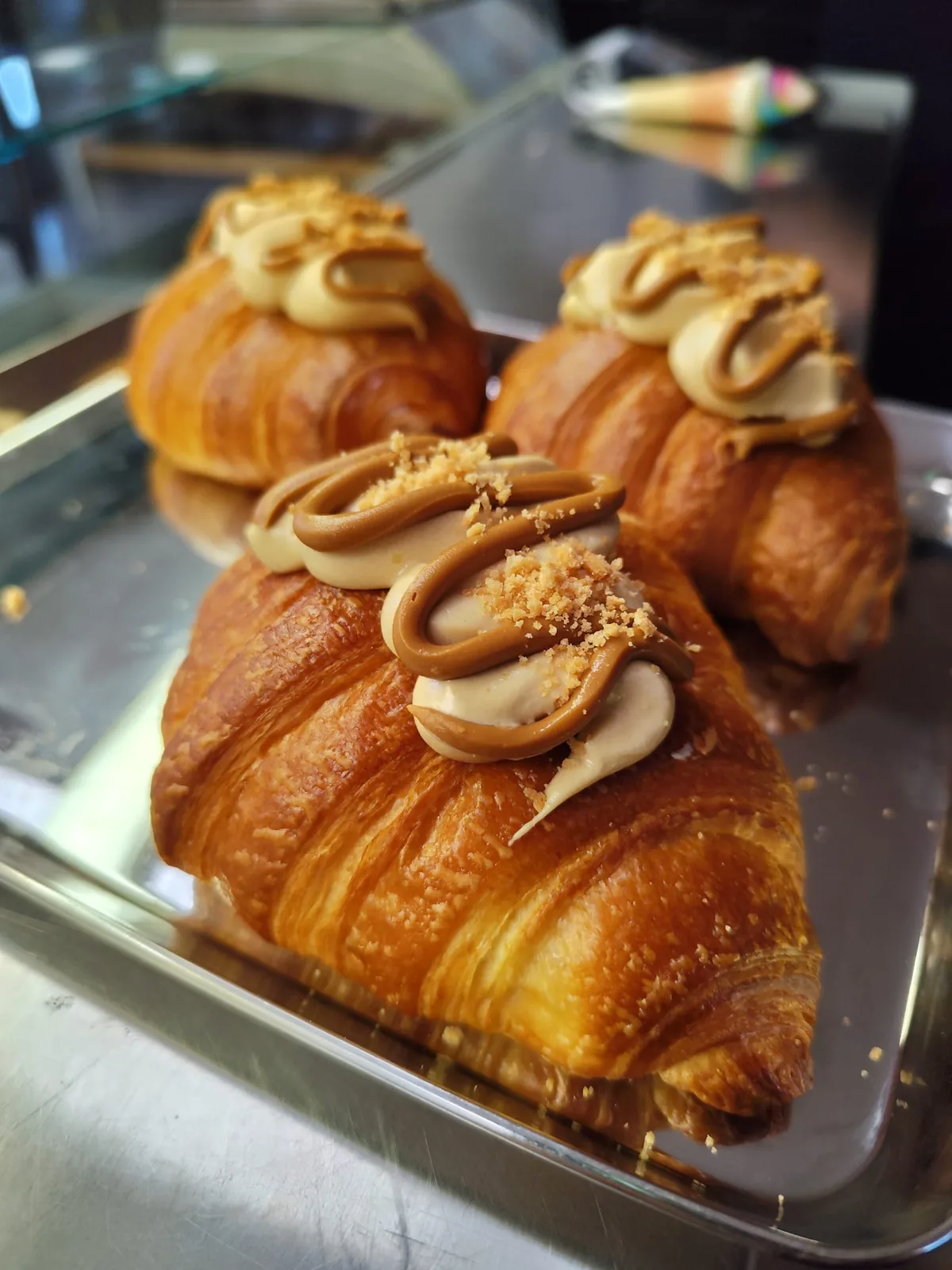 Croissant farciti con crema al caramello e granella, disposti su una teglia dietro il vetro della vetrina.