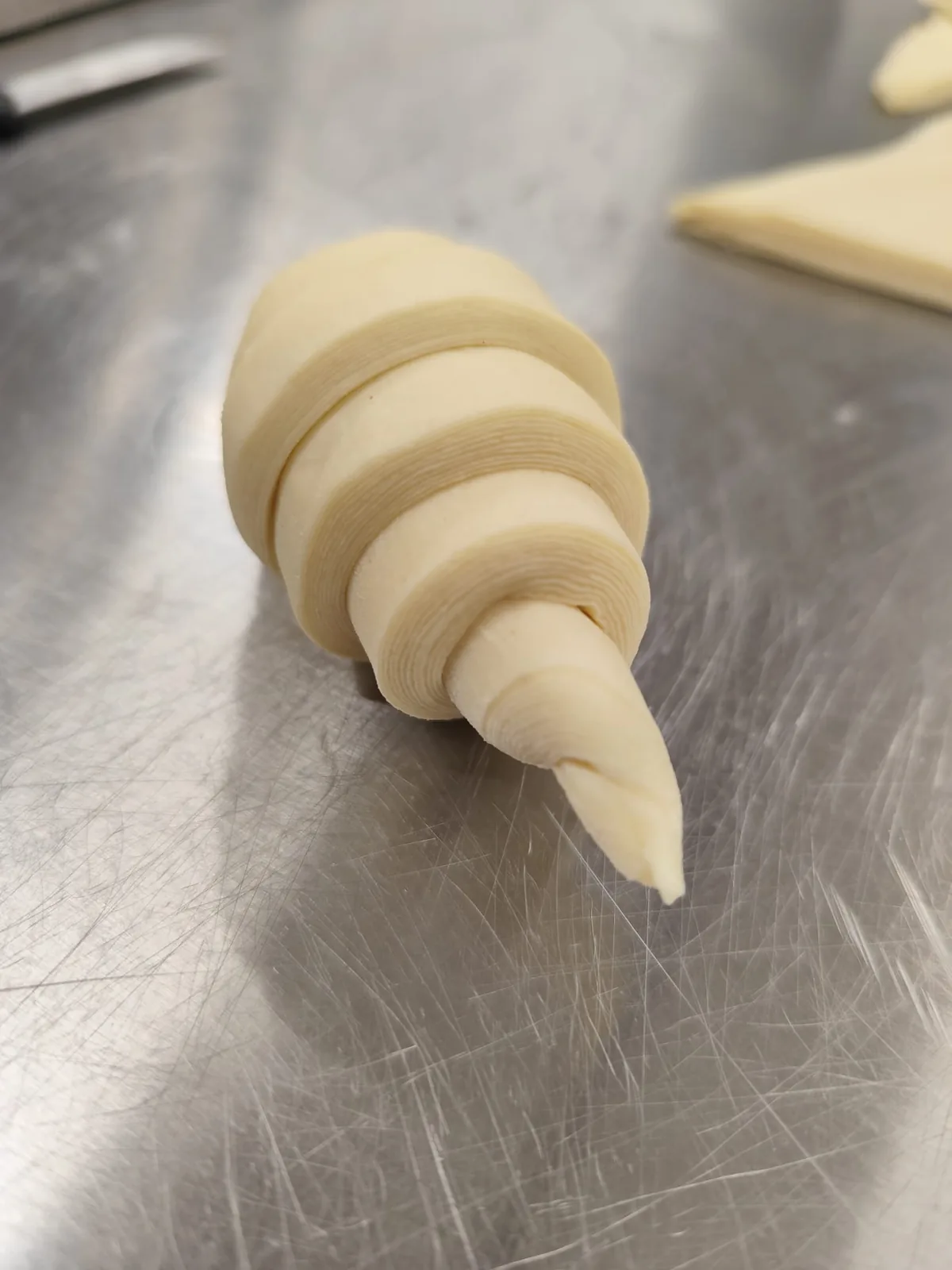 Un croissant arrotolato a mano, ancora crudo, sul banco d'acciaio del laboratorio: sulla punta si contano gli strati di sfoglia.
