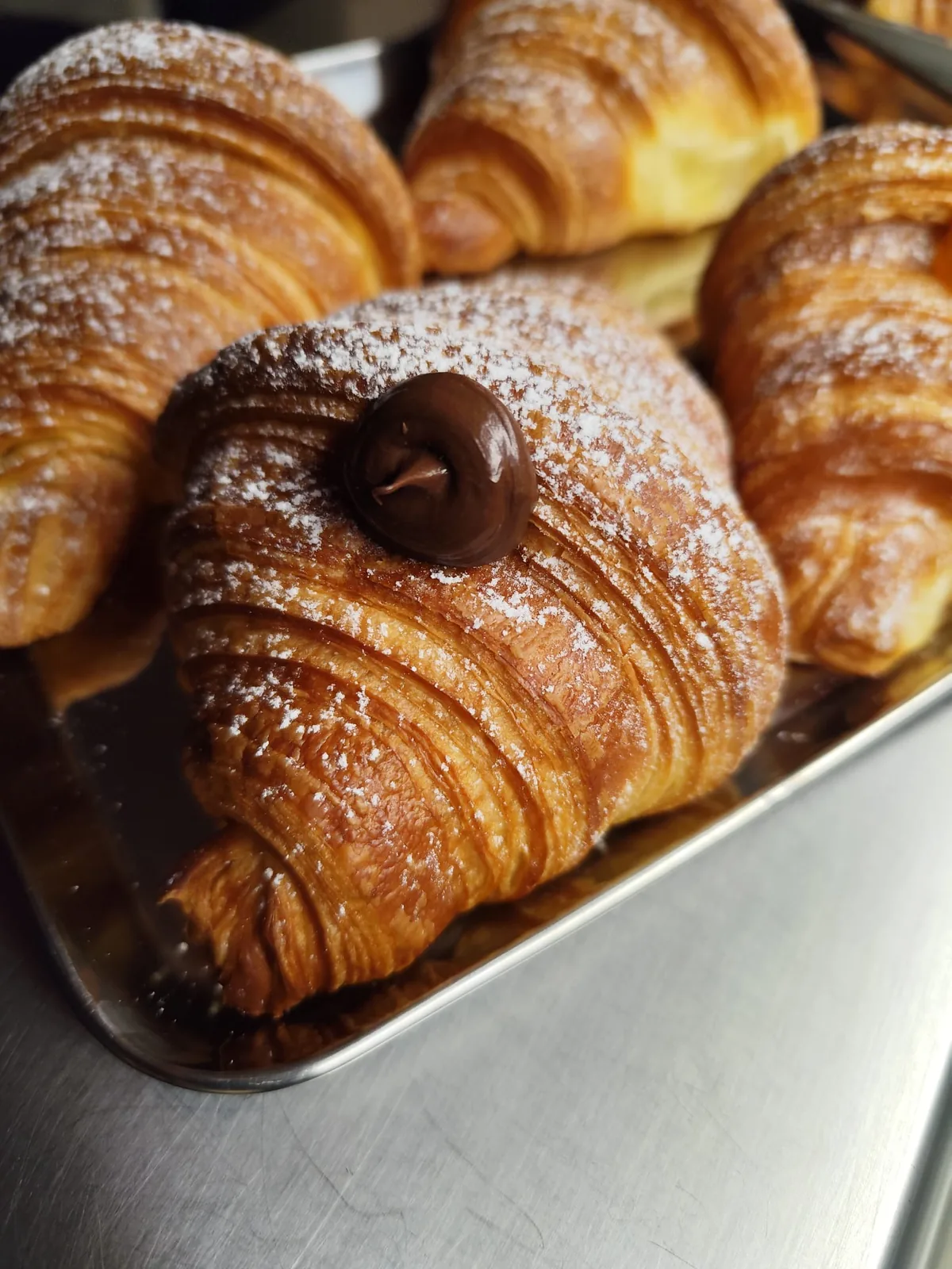 Croissant sfogliati spolverati di zucchero a velo, con una goccia di crema al cioccolato, allineati su una teglia d'acciaio.