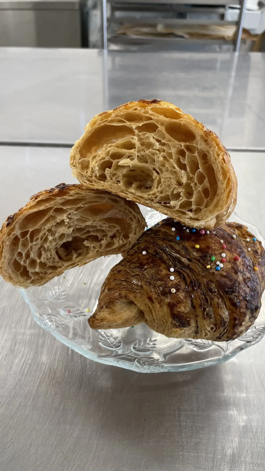 Un CroisSard tagliato a metà: l'alveolatura della sfoglia in sezione, accanto a un croissant intero su un piatto di vetro.