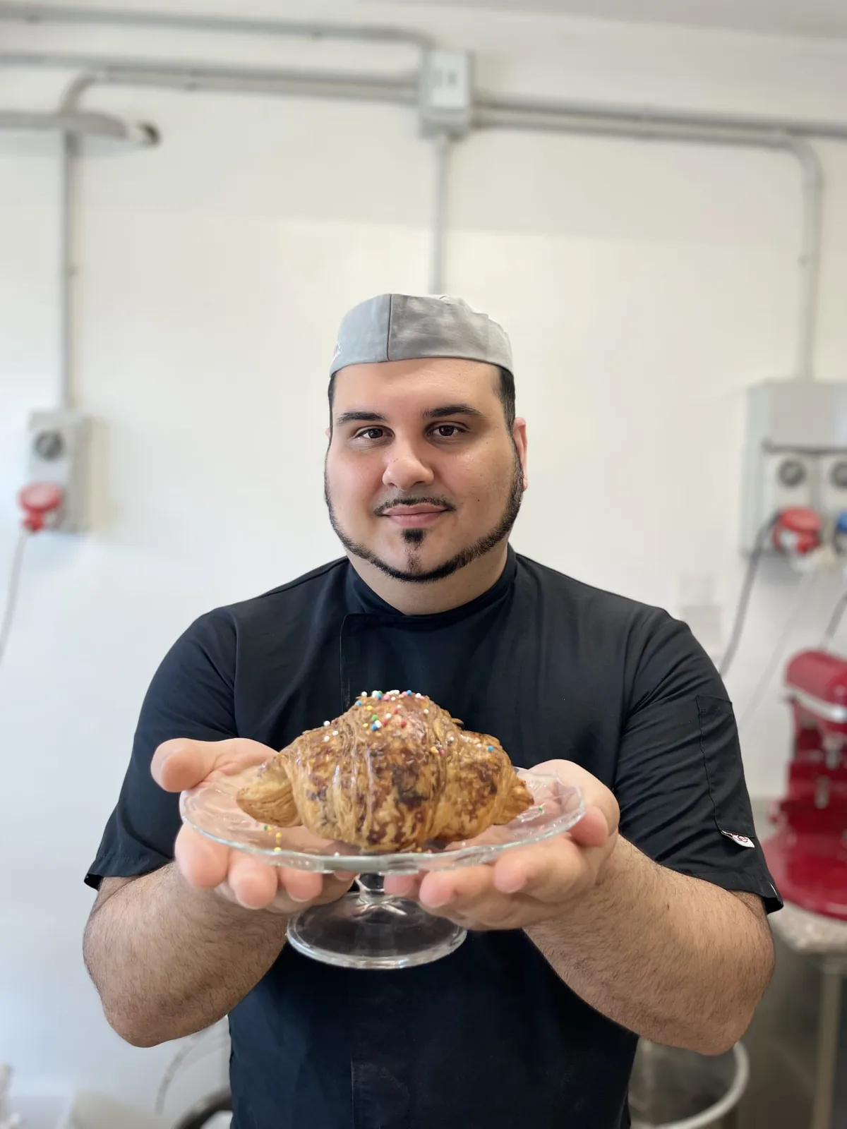 Nicola Cara in laboratorio, con la giacca nera e il copricapo, mentre presenta un CroisSard su un'alzatina di vetro.