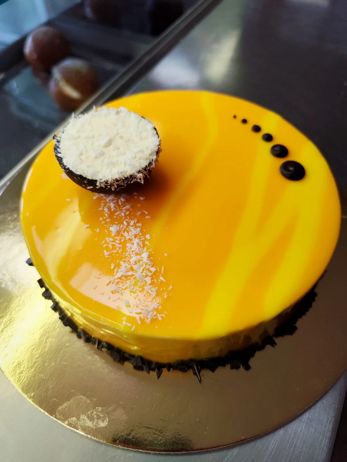 Torta moderna con glassa a specchio gialla, decorata con cocco grattugiato, gocce di cioccolato fondente e una semisfera bianca.