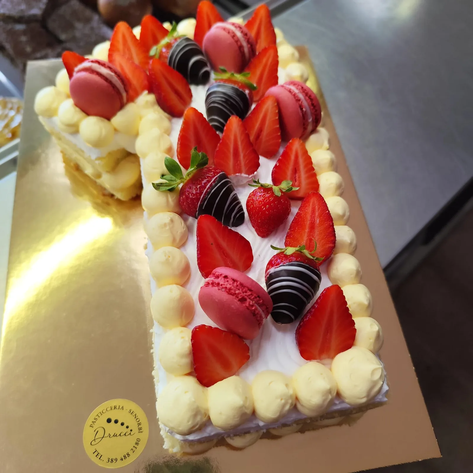 Torta rettangolare alla crema e fragole, decorata con macaron rosa e fragole ricoperte di cioccolato.